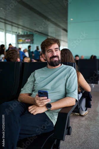 Smiling man sitting using phone while waiting for his flight at the airport departure area, looking the views. Vertical image with copy space.