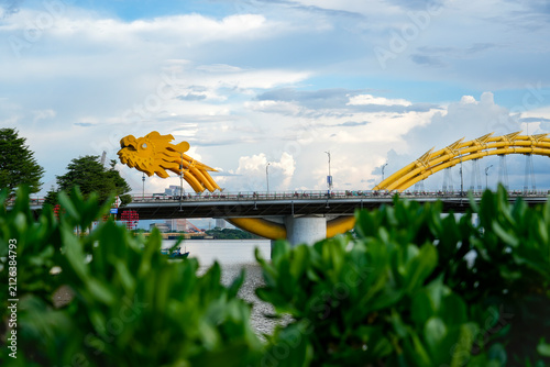 The striking head golden dragon head of Da Nang's Dragon Bridge stands over the Han River, flanked by green trees and decorative red lanterns under a cloudy sky