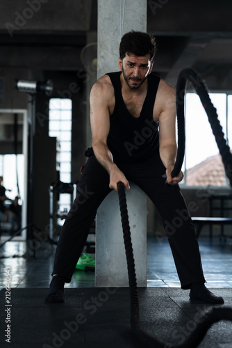 Fotografie Adult Caucasian man gripping battle rope in modern gym, male athlete pushing arm