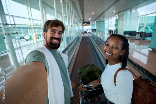 Young diverse couple smiling and taking a selfie on an airport moving walkway, looking at camera cheerfully.