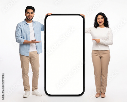 Young Indian couple standing beside a giant blank smartphone screen while presenting a mobile app concept with cheerful expressions isolated on a white background for commercial design