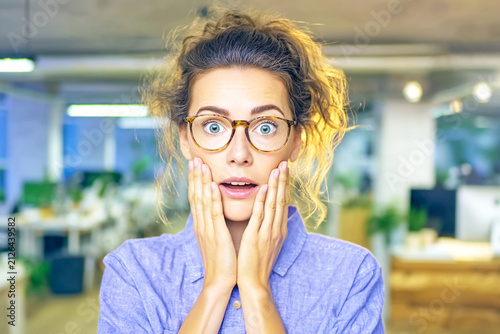 A young professional woman with messy hair and glasses expresses a look of surprise and shock while holding her hands to her face in a modern office, capturing a moment of genuine human