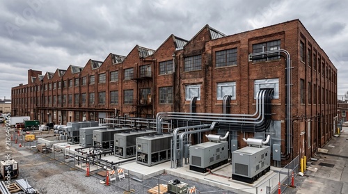 Elevated View of a Long Red Brick Sawtooth Roof Factory Retrofitted With Modern Air Cooled Chillers Generator Enclosures and Cable Bundles Curving Up the Facade Adaptive Reuse Industry