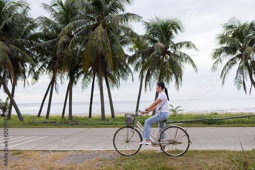 Konstfotografi Asian Woman Cycling on Coastal Road Lined with Coconut Trees,A candid, full-body photograph of a young Asian woman in her late 20s riding a vintage cruiser bicycle with a front basket along a quiet