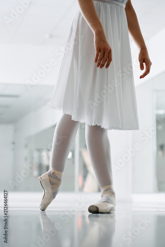 Fotografie Cropped low-angle view of a female ballet dancer in a white dress and point shoes practicing elegant footwork in a bright modern dance studio