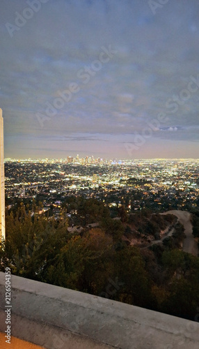 The illuminated cityscape of downtown Los Angeles at dusk, framed by the dark hillside trees of Griffith Park and the concrete terrace under a dramatic cloudy evening sky. USA.
