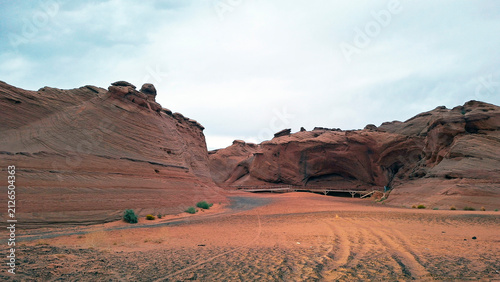 Eroded sandstone canyon walls and layered rock formations. Desert of Page, Arizona, featuring sandy soil with tire tracks and a distant wooden pedestrian bridge spanning across the rugged terrain.