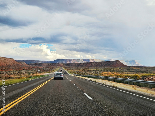 Two-lane asphalt highway with double yellow lines,  cars driving towards distant geological rock formations, and desert mesas near Page, Arizona, under heavy overcast and white cumulus storm clouds. 