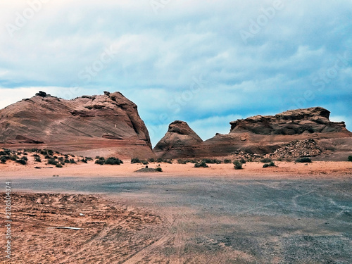 Sandstone buttes and eroded rocks in the desert near Page, Arizona, with sparse desert shrubs on the sandy soil under a dramatic and heavy overcast blue sky. USA