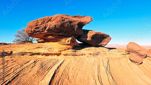 Geological formation of red sandstone balanced rocks showing prominent sedimentary layers, surrounded by a barren desert floor and a small green bush near Page, Arizona, under a vibrant blue sky. USA