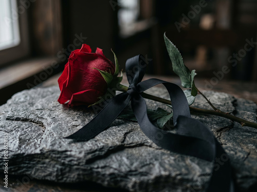 Red rose flower tied with black mourning ribbon on dark stone