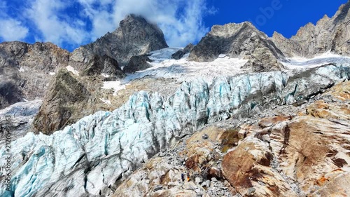 Aerial drone view of the Frébouge glacier in the Mont Blanc massif near Courmayeur, Italy.  Hikers on the rocky terrain near the ice glacier showcase the massive scale of the natural landscape. 