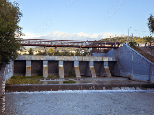 Urban Flood Control Dam with Pedestrian Bridge and Flowing River at SunsetA modern flood control dam with cascading water, a pedestrian bridge, and riverside infrastructure surrounded by lush trees un