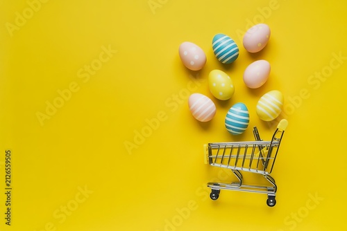 Flat lay of colorful easter eggs spilling from a miniature shopping cart on a vibrant yellow background