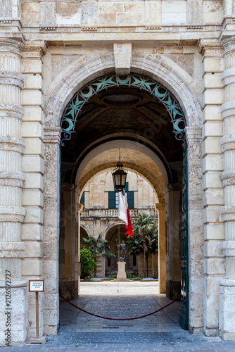 A baroque stone archway with star-and-crescent ironwork opens onto the Auberge de Castille courtyard, framing a Neptune statue and Maltese flag