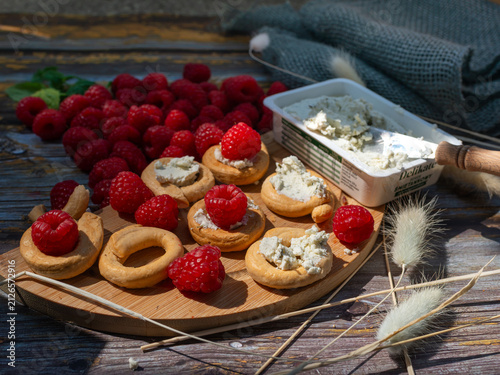 Foto Crisp ring-shaped crackers topped with creamy herb cheese and ripe raspberries, Rubus idaeus, beside an open cheese container and spreading knife