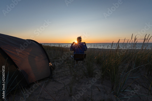 Canvastavla Solitary traveler sitting beside a tent on the sandy dunes of Nõva beach in Estonia, watching a dramatic Baltic Sea sunset with sunstar flare