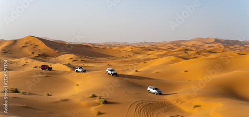 Off-road vehicles drive across vast sand dunes in the Sahara Desert, Morocco. They create tracks in the sand under the bright sky during daylight hours.