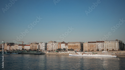 Photography A river cruise ship glides past Budapest's historic Danube-side buildings, their facades bathed in warm afternoon light beneath a clear blue sky along the calm waterfront