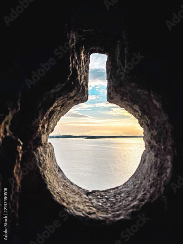 Bild auf Leinwand Scenic sea view through a medieval stone loophole with warm sunset light reflecting on calm water