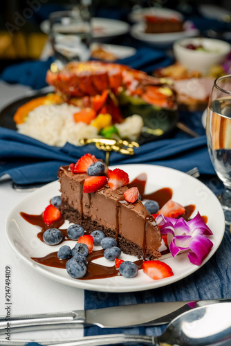 Yummy dessert on the table. A chocolate cake slice is on a white plate, with fruits and sauce around it, during a special dinner in a restaurant.