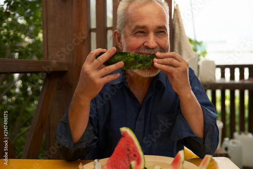 Portrait of an adult man eating a slice of watermelon on the terrace of a summer house.
