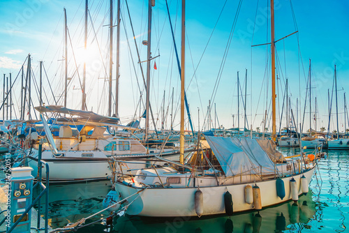 Sailing boats with tall masts are moored in a marina under bright blue sky.