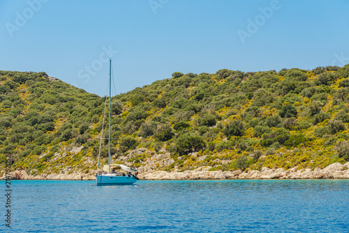 White sailing yacht anchors near a lush green rocky hillside above turquoise sea under a clear blue sky