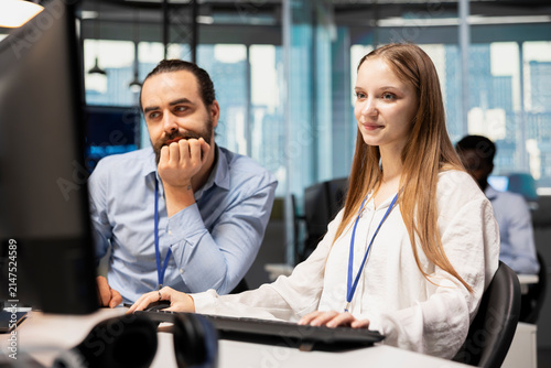 Senior team leader mentors a junior developer at an office desk and teaches coding best practices, guiding the female intern through technology company project tasks during workplace training.