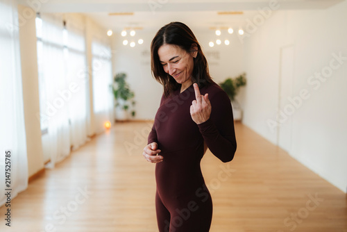 Middle-aged woman making an obscene gesture in a sunlit yoga studio, realistic photography, warm background, defiance concept