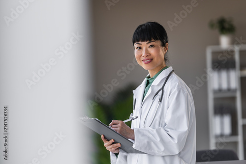 Canvastavla Female doctor with stethoscope smiling while taking notes on clipboard in modern