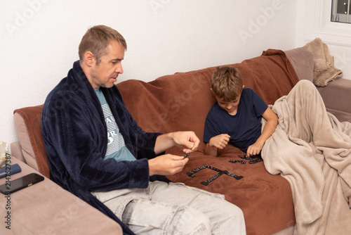 Father and son play dominoes together on a couch in their living room during the evening