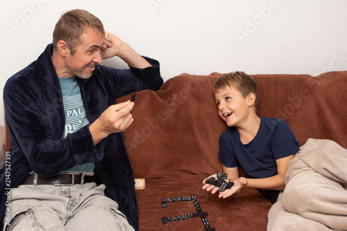 Father and son play dominoes on a couch in a living room at home during the evening