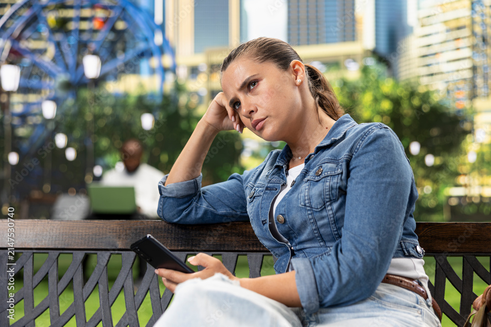 Fototapeta premium Thoughtful woman rests her head on one hand while holding a smartphone on a park bench, taking a reflective pause after a disappointing message.