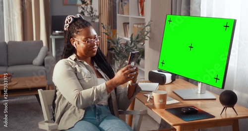 Smiling young Black woman uses her smartphone while sitting at a computer in the living room, with a green screen and tracking markers visible on the monitor.