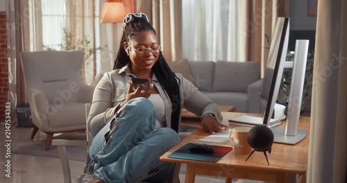 Young Black woman works remotely from home, listening and asking an AI assistant on her smartphone while typing on her computer in her cozy living room.
