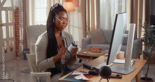 Young Black woman patient holds pills and carefully listens to doctor’s advice during a medical video call at home in her living room.