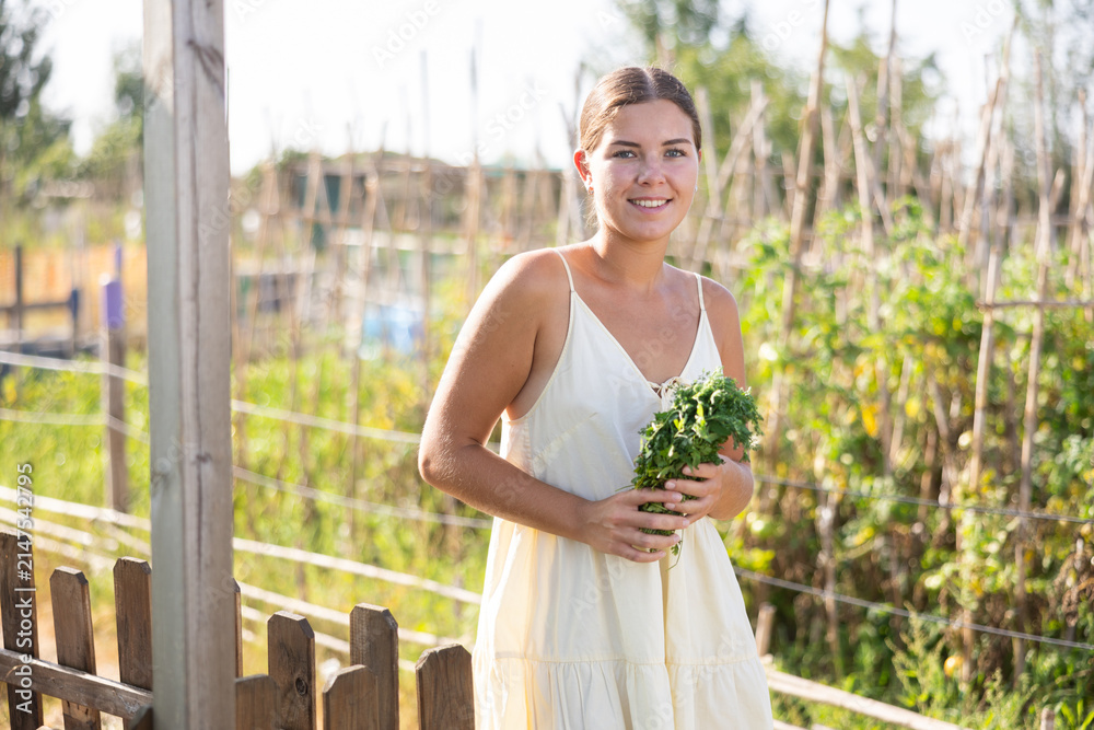 Obraz premium European girl stopped near bed of tomatoes, utility shed and barn, takes break from gardening, enjoys active hobby of growing garden crops. Girl with agricultural tool in hands, stands near trellis