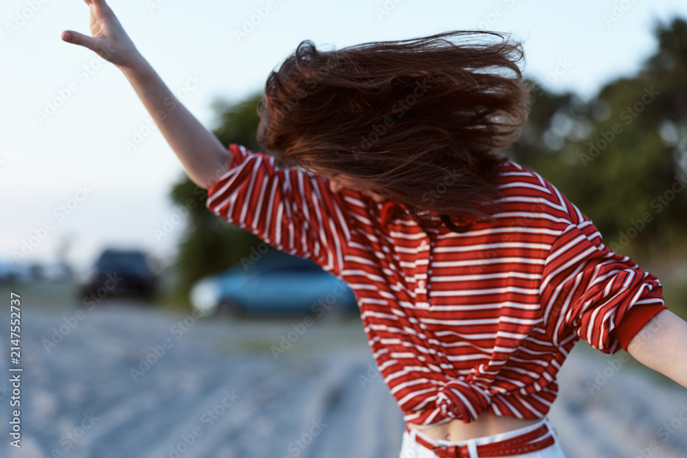 Fototapeta premium Young woman in a striped red shirt raises her arms in joy on a breezy beach path, capturing a spontaneous carefree lifestyle moment.