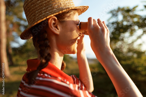 Canvas Print A young girl in a striped shirt and straw hat peers through binoculars in a sunl