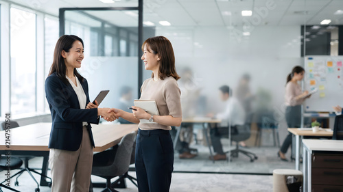 オフィスでノートやタブレットを手に向かい合い笑顔で握手する女性社員たち