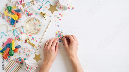 Party preparation: Hands arranging colorful confetti, balloons, streamers, and party hats, with a glass of champagne in a festive setting