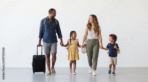 A smiling family of four, holding hands, with two children and parents, are walking with luggage in a bright, modern space. The father is pulling a suitcase, and the mother carries a handbag.