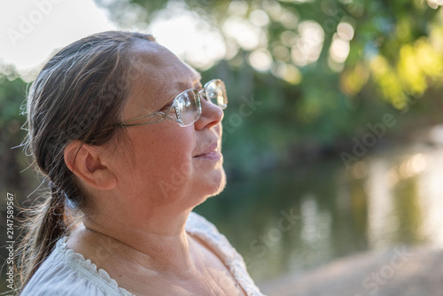 Happy middle-aged woman with glasses, mental health issues, outdoors relaxation, authentic reaction, emotional season Mature portrait, golden hour light, peaceful expression, blurred nature background