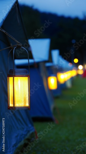 At dusk, the camping tents on the green grass are illuminated by local industrial outdoor lights, with a close-up shot of the warm light.