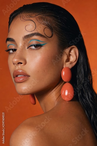 Stylish young woman with bold geometric face art and vibrant coral statement earrings, glamorous portrait against warm orange gradient background, modern makeup and fashion concept