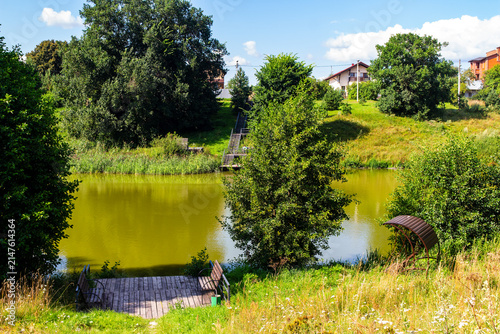 Fotografía Serene countryside pond featuring a rustic wooden deck with benches, surrounded