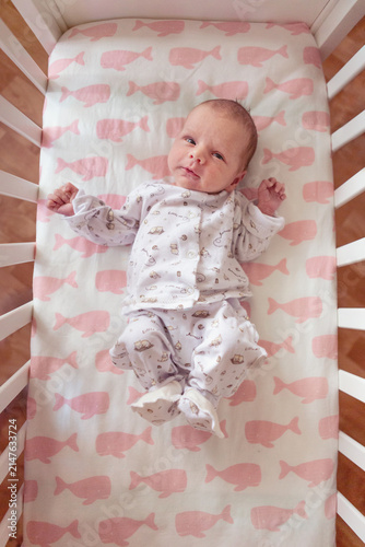 A cute newborn baby lies awake in a white crib on a soft pink whale patterned sheet, wearing cozy cotton pajamas and looking curiously at the camera in a warm, peaceful nursery filled with light.