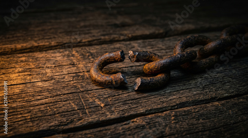 Broken rusty iron chain link resting upon dark weathered timber planks under dim warm lighting with separated dark textured textured wooden surface single fractured heavy