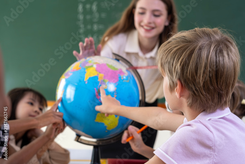 Smiling female teacher guiding elementary students with a world globe. Curious schoolboy pointing at continents on globe during geography class. Young diverse students learning world geography.